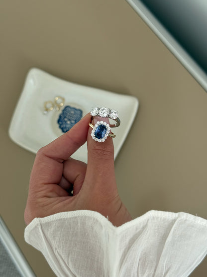 A close-up of a hand wearing a ring with a large blue sapphire surrounded by a halo of small clear stones, set on a gold band. 