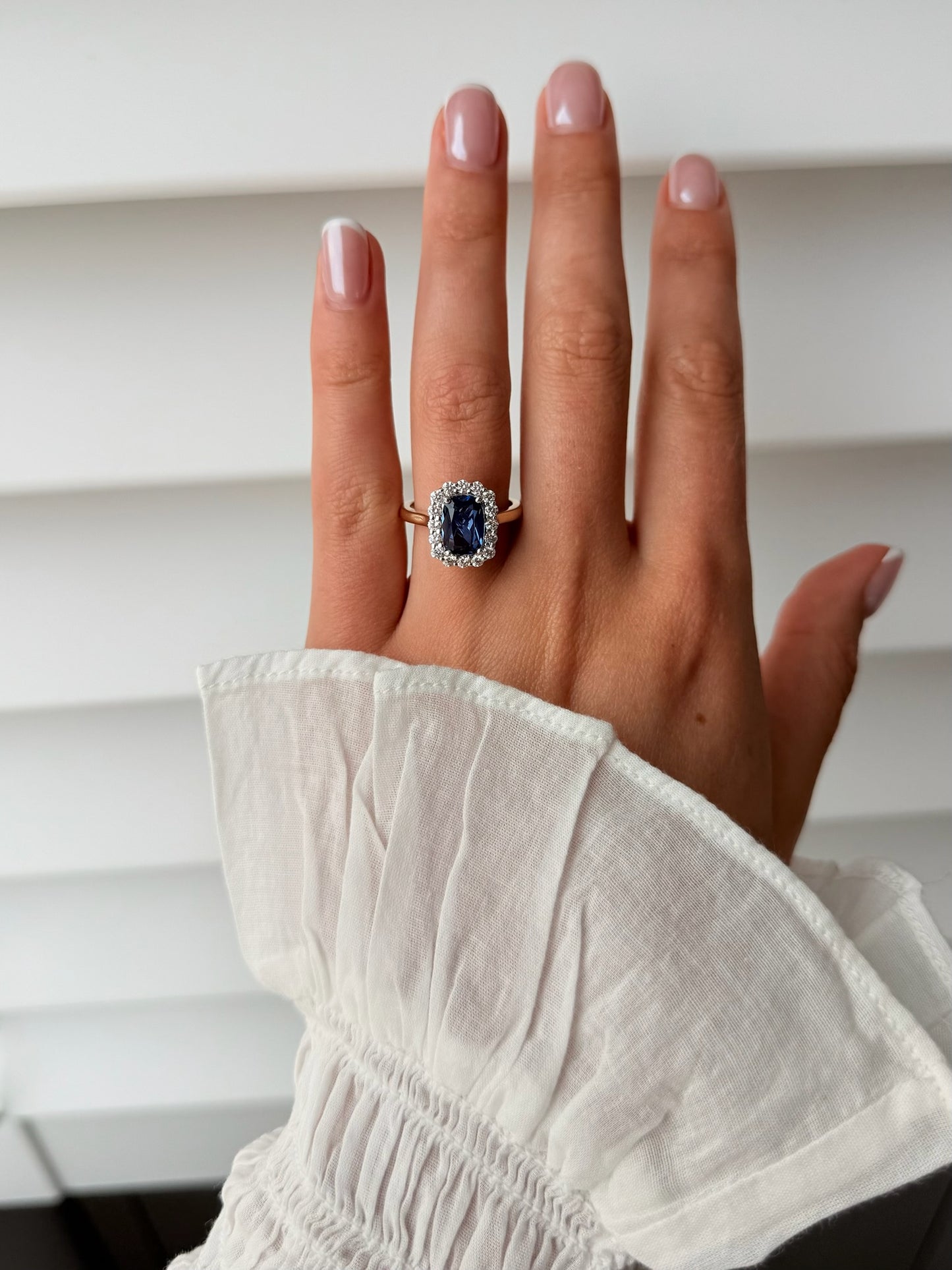 A close-up of a hand wearing a ring with a large blue sapphire surrounded by a halo of small clear stones, set on a gold band. 