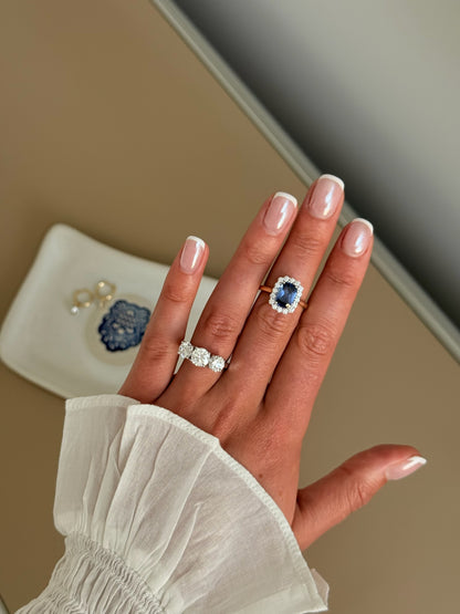 A close-up of a hand wearing a ring with a large blue sapphire surrounded by a halo of small clear stones, set on a gold band. 
