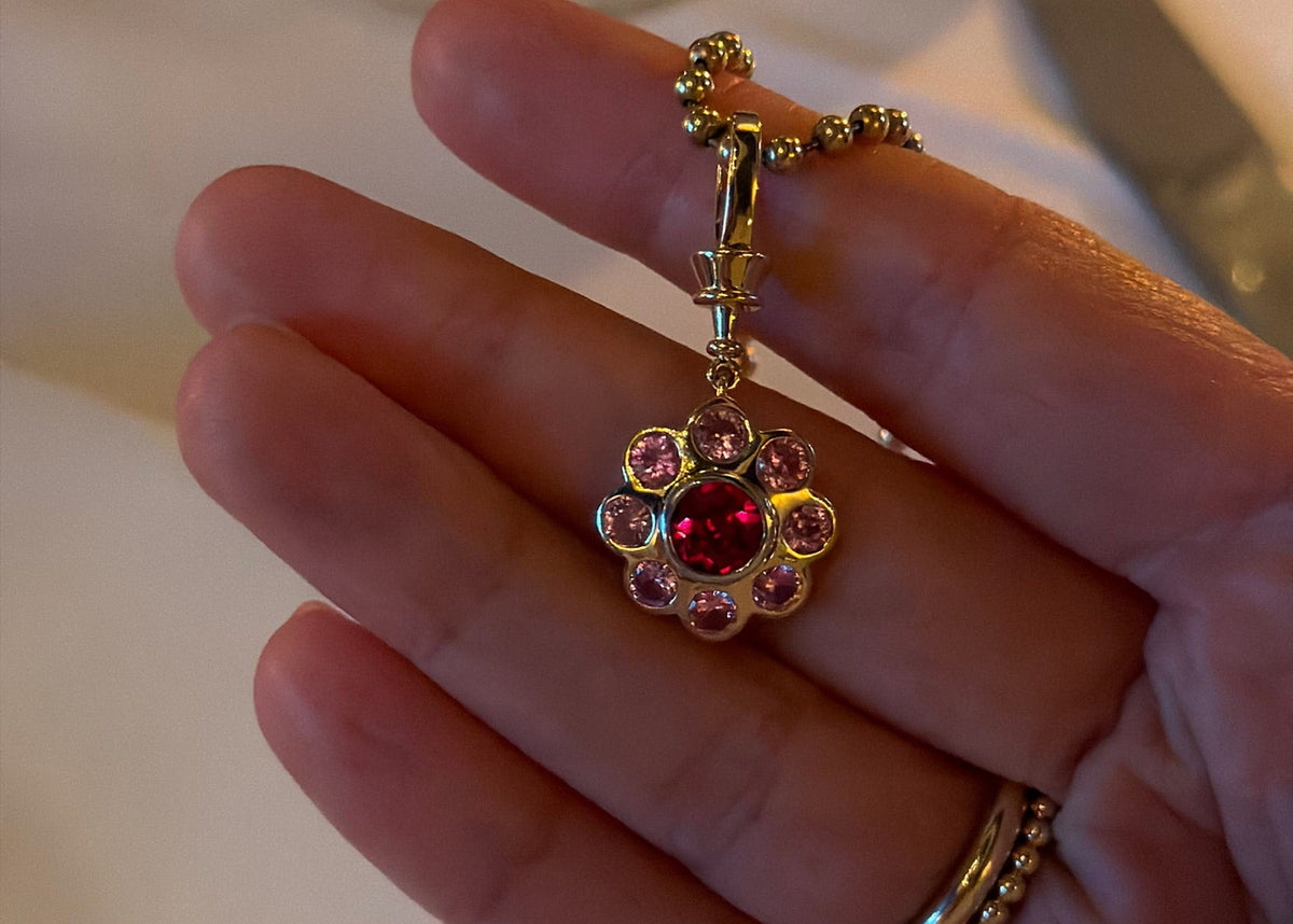 A gold-tone flower-shaped pendant charm with a central red gemstone and surrounding pink stones, held in a person's hand over a softly blurred dining table background.