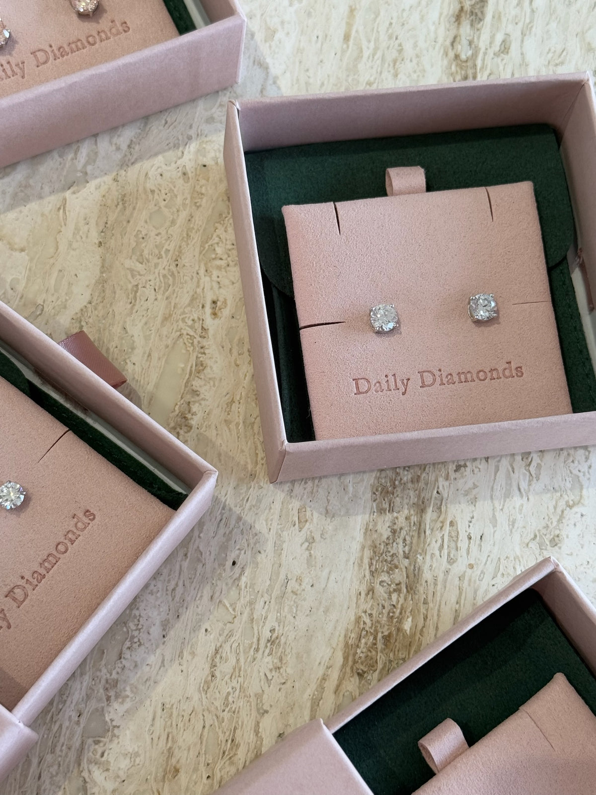 Pair of diamond stud earrings in a 'Daily Diamonds' box on a textured surface.