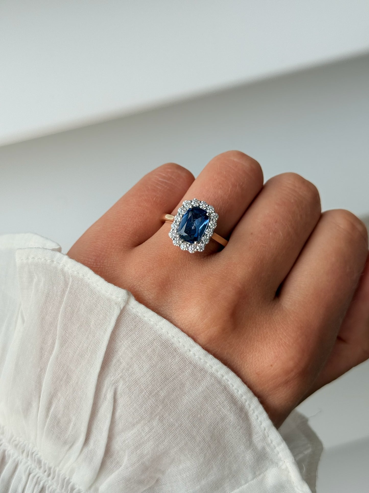 A close-up of a hand wearing a ring with a large blue sapphire surrounded by a halo of small clear stones, set on a gold band. 