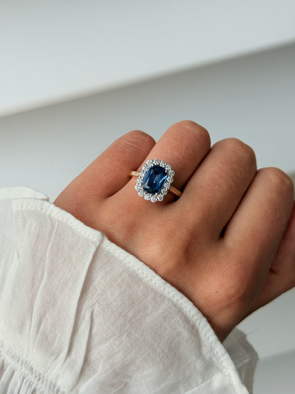 A close-up of a hand wearing a ring with a large blue sapphire surrounded by a halo of small clear stones, set on a gold band. 