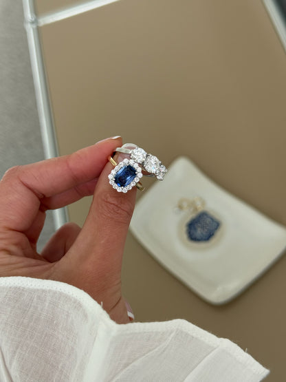 A close-up of a hand wearing a ring with a large blue sapphire surrounded by a halo of small diamonds, set on a gold band. 