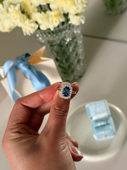A close-up of a hand wearing a ring with a large blue sapphire surrounded by a halo of small clear stones, set on a gold band. 