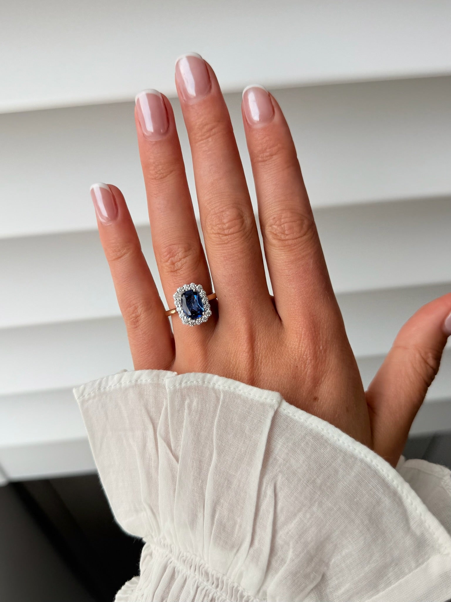 A close-up of a hand wearing a ring with a large blue sapphire surrounded by a halo of small clear stones, set on a gold band. 