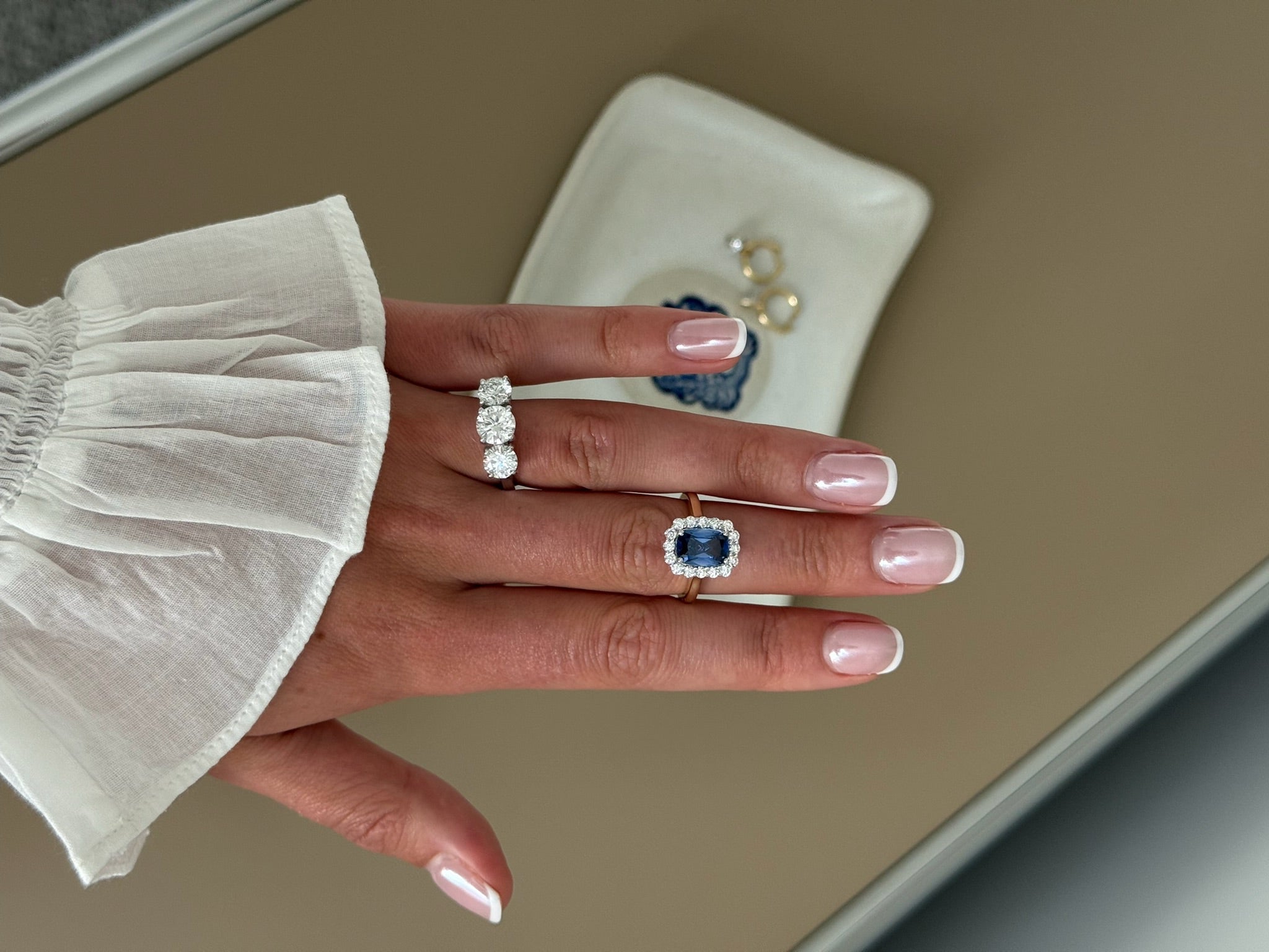 A close-up of a hand wearing a ring with a large blue sapphire surrounded by a halo of small clear stones, set on a gold band. 