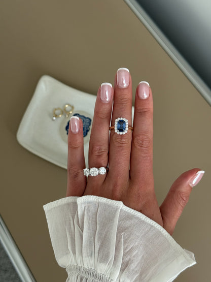 A close-up of a hand wearing a ring with a large blue sapphire surrounded by a halo of small clear stones, set on a gold band. 