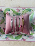 Four slim identity bracelets displayed on a pink velvet pillow, resting on a floral-patterned tray. The bracelets include silver and gold metals with heart and flower charms.