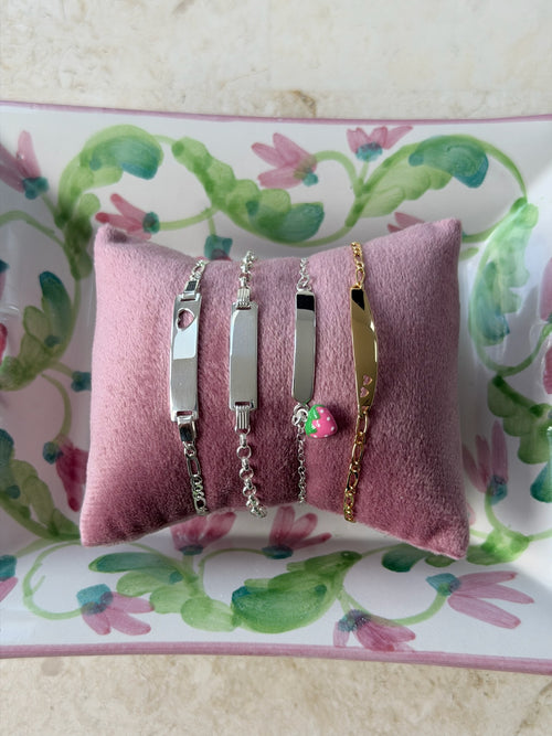 Four slim identity bracelets displayed on a pink velvet pillow, resting on a floral-patterned tray. The bracelets include silver and gold metals with heart and flower charms.