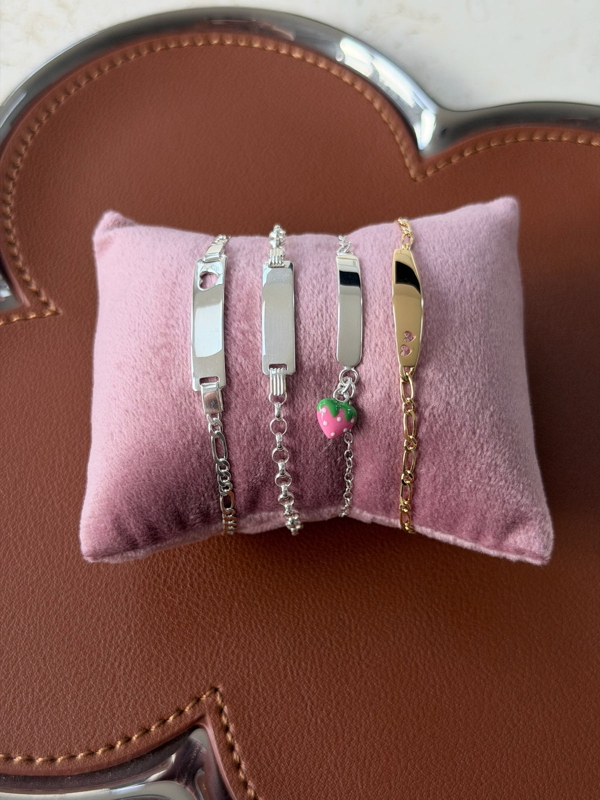 Four personalized ID bracelets displayed on a pink velvet cushion, set against a brown textured leather background. The bracelets include silver and gold metal plates, chain links, and a small pink strawberry charm.