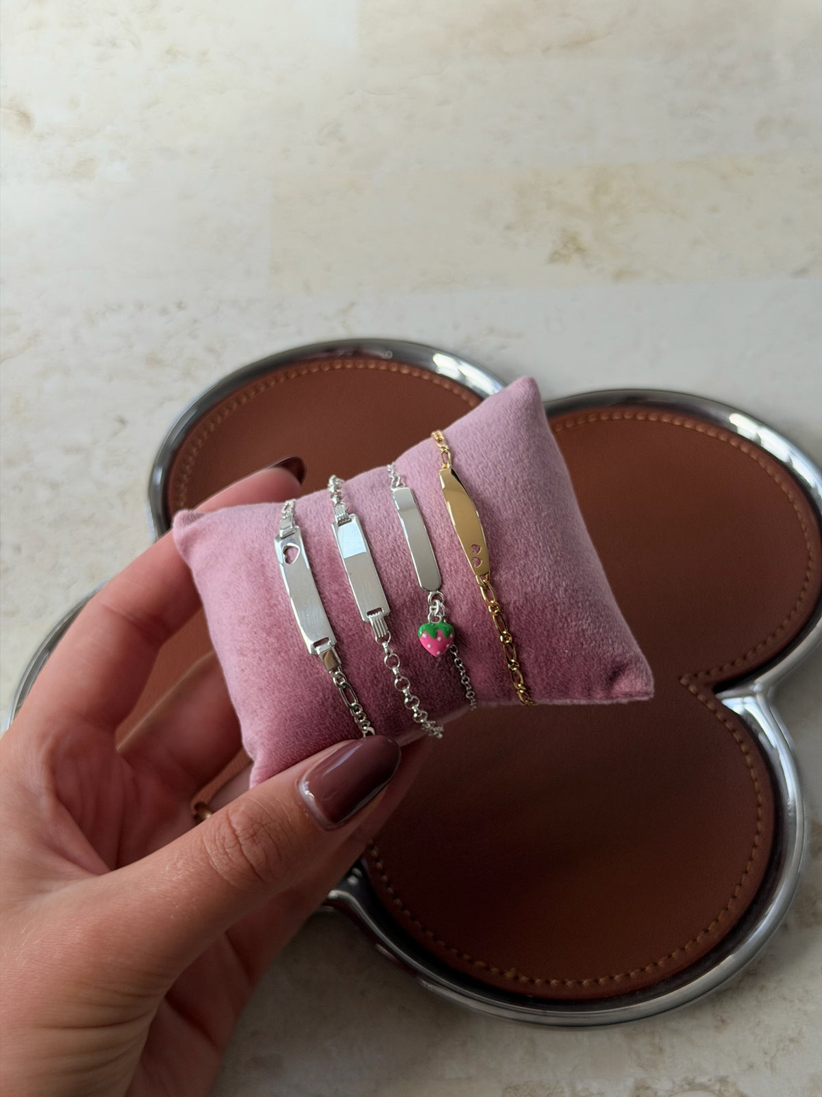 A hand holds a small pink velvet jewelry display cushion with four bar-style bracelets attached. The bracelets are silver and gold-toned, with one small pink heart charm.